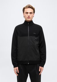 Jeune homme debout face à l'avant, portant une veste noire à fermeture éclair avec un col montant et le logo "BOSS" sur la poitrine.