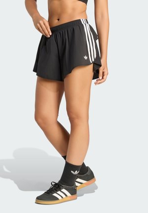 Person iført sorte atletikshorts med hvide striber, sorte Adidas sokker og sorte Adidas sneakers, der står med det ene ben over det andet.
