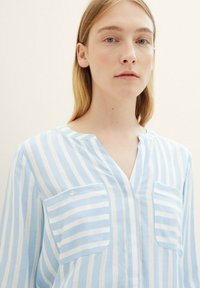 Camisa a rayas azul claro y blanca con escote en V, dos bolsillos en el pecho y un corte relajado. La tela parece ligera y transpirable.