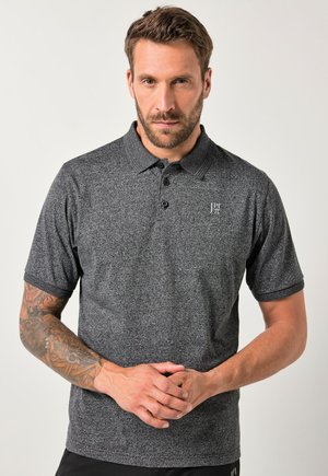 Mand med kort brunt hår og skæg, iført en grå kortærmet polo-shirt med tatoveringer på venstre arm, stående foran en ensfarvet baggrund.