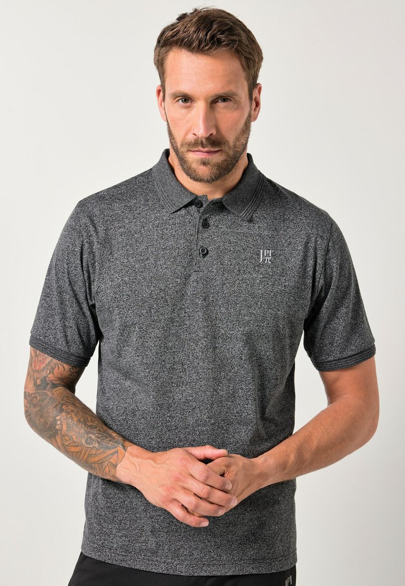 Uomo con capelli corti e marroni e barba, che indossa una polo grigia a maniche corte con tatuaggi sul braccio sinistro, in piedi su uno sfondo semplice.