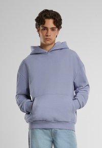 Lyselilla hettegenser laget av myk stoff, med en frontlomme, snorhette og avslappet passform. Båret med lyseblå jeans.