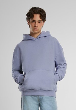 Lyselilla hettegenser laget av myk stoff, med en frontlomme, snorhette og avslappet passform. Båret med lyseblå jeans.