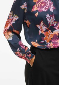 Blusa floreale con sfondo blu navy, fiori vibranti rosa e arancioni, e polsini elasticizzati, abbinata a pantaloni neri.