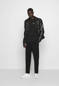 Giubbotto piumino nero con una texture lucida, t-shirt nera con logo, pantaloni neri e sneakers bianche, su uno sfondo neutro.