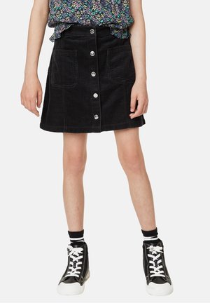 Pige iført en sort kordfløjlsnederdel med knapper foran og store lommer, blomsterprintet top, sorte ankelsokker og sorte høje sneakers med hvide snørebånd.