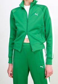 Veste verte zippée avec des accents blancs, assortie à un pantalon. Fabriquée en tissu lisse, avec un logo et des poches zippées.