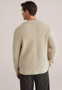 Pulover tricotat texturat bej, cu guler larg și umeri căzuți, asociat cu pantaloni închisi la culoare. Designul prezintă un tiv și manșete cu model smoca.