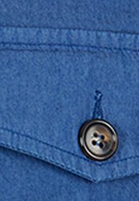Tessuto in denim blu con colletto a punta e un bottone nero a quattro fori, dettagli cuciti visibili sulla superficie del tessuto.