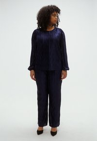 Σετ ρούχων σε σκούρο μπλε χρώμα με πιέτες, αποτελούμενο από μπλούζα με μακριά μανίκια και φαρδιά παντελόνια. Κατασκευασμένο από απαλό ύφασμα με ελαφριά γυαλάδα.