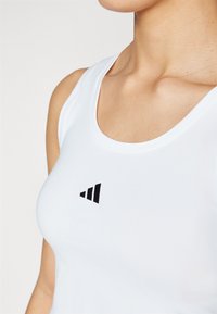 Top atlético branco com um logotipo preto na forma de triângulos empilhados. O tecido é suave e ajustado, com decote em forma de scoop.