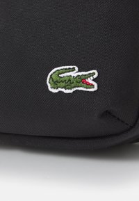 Black fabric bag featuring a green embroidered crocodile logo with red accents, positioned in the lower corner against a textured background.