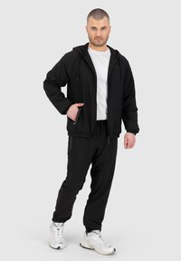 Giacca nera con zip e cappuccio, abbinata a pantaloni elastici neri e t-shirt bianca. Sneakers bianche. Tessuto liscio, design a vestibilità moderna.