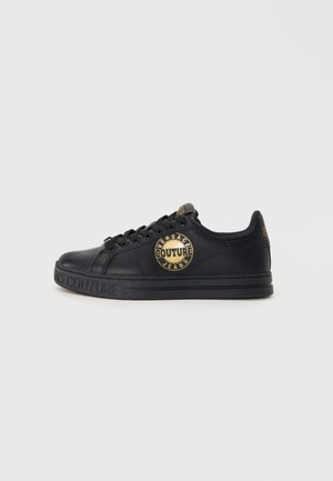 Sneakers en cuir noir avec un logo rond portant "Versace Couture" en or. Design élégant, semelle plate et surfaces texturées.