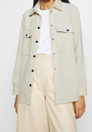 Osoba ubrana w beżową kurtkę z sztruksu zapinaną na guziki, na białej koszulce i kremowych szerokich spodniach z wysokim stanem, pokazana od szyi do połowy uda.