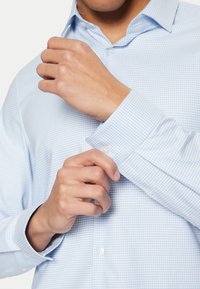 Camisa de cuadros azul claro, hecha de algodón; presenta un cuello abotonado y botones de plástico transparente. Los puños están ligeramente arremangados.