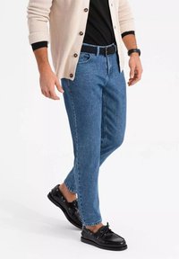 Vaqueros de denim azul con un corte recto, llevados con un cinturón negro y combinados con zapatos negros. El conjunto está complementado con un cárdigan crema.