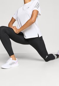 Weißes Sport-T-Shirt mit grauen Schulterstreifen, schwarze Leggings und weiße Sneakers mit rosafarbenen Akzenten, verfügen über eine schlanke Passform und seitliche Schlitze.