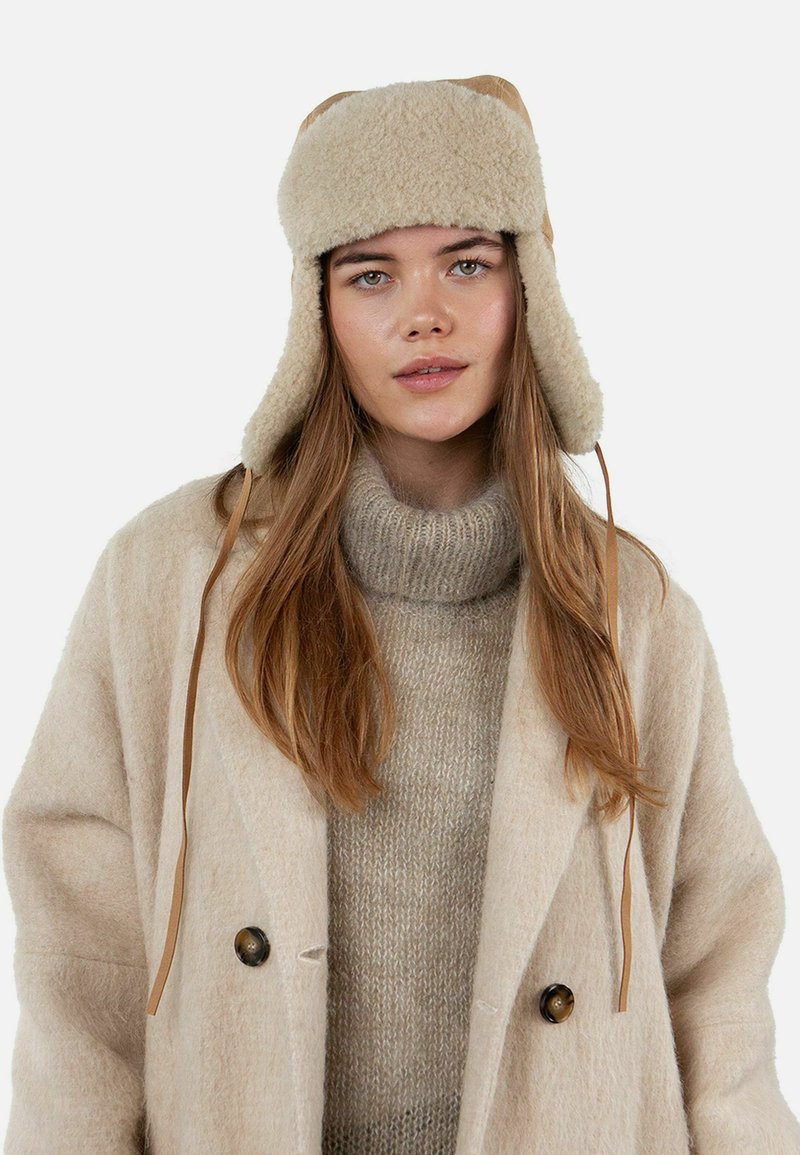 Beige fur hat with ear flaps, featuring tie straps. The model wears a soft beige sweater and an oversized light beige coat.