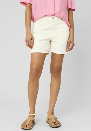 Femme portant un short en jean blanc, une chemise rose à manches courtes avec boutons, des sandales beiges et un bracelet de cheville blanc en coquillage, debout devant un fond clair.