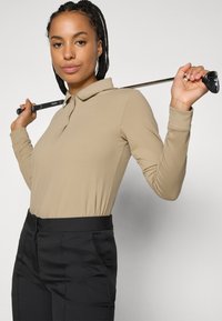 Beige pitkähihaisessa polo-paidassa, jossa on kaulus, yhdistettynä mustiin housuihin. Malli pitää golfmailaa, esitellen urheilullista pukeutumista.