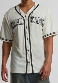 Kremowa koszula z krótkim rękawem z czarnymi akcentami, dekoltem w serek i guzikami. Na piersi znajduje się napis "KARL KANI" w pogrubionych literach.