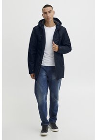 Navy waterproof jacket with a hood, front zip, and pockets; paired with blue jeans and black sneakers. Smooth fabric texture.