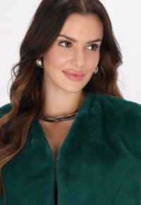 Emeraldgrøn faux fur-frakke med en blød tekstur, der har en V-udskæring og et sølvfarvet lagdelt halskæde, der fremhæver udskæringen.