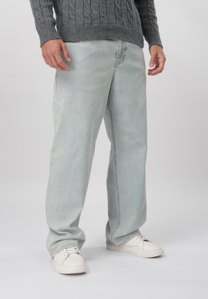 Homme vêtu d’un jean bleu clair ample, d’un pull gris torsadé et de baskets blanches, debout devant un fond uni.