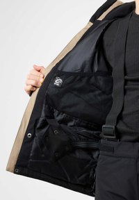 Beige und schwarze Jacke mit einer Netz-Innentasche, verstellbaren Riemen und Druckknöpfen. Das Innenfutter ist mit einem glatten, glänzenden Stoff ausgekleidet.