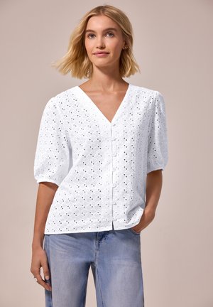 Kvinde med kort blondt hår iført en hvid bluse med øjer og lyseblå jeans, poserer med den ene hånd i lommen.