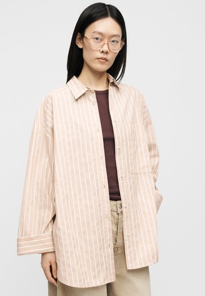 Femme aux longs cheveux noirs et lunettes portant une chemise oversize beige et blanche rayée sur un haut marron et un pantalon beige, debout devant un fond gris.