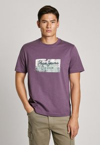 Violetinės spalvos medvilninis marškinėlis su centrinėmis grafikos spaudomis, kuriuose užrašas "Pepe Jeans" ir tekstūruotas fonas. Apvalus kaklas ir trumpomis rankovėmis.