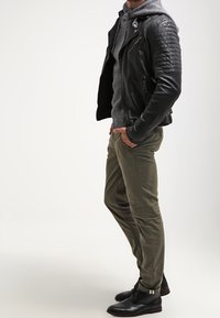 Mężczyzna stojący z rękami w kieszeniach, ubrany w pikowaną czarną skórzaną kurtkę, szarą bluzę z kapturem, oliwkowe spodnie i czarne eleganckie buty na jednolitym tle.