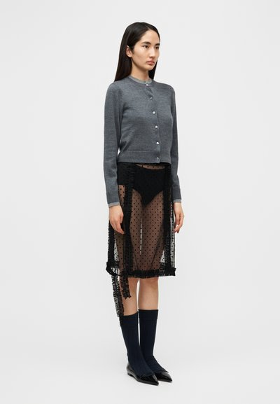 Γκρι ζακέτα με στρογγυλή λαιμόκοψη και κουμπιά, συνδυασμένη με διαφανή μαύρη φούστα πουά με βολάν, καλτσάκια μέχρι το γόνατο και μυτερές μπαλαρίνες.