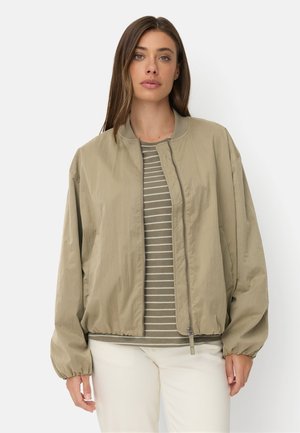 Femme portant une veste bomber marron clair zippée sur une chemise rayée verte et blanche et un pantalon blanc, debout devant un fond blanc uni.