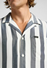 Chemise à manches courtes rayée en gris et blanc, avec une poche poitrine et des boutons blancs. Tissu doux avec une texture légère.