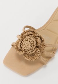 Beige sandal with a square toe and a woven flower detail on the upper. Straps are thin and match the color of the sole.
