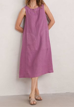 Frau trägt ärmelloses, locker sitzendes violettes Kleid und beige Sandalen, steht mit den Händen hinter dem Rücken vor einer schlichten Wand.
