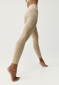 Beżowe legginsy o smukłym kroju, z złotymi akcentami przy szwach i wysokim stanem. Gładka faktura, dopasowany kształt i elastyczny materiał.