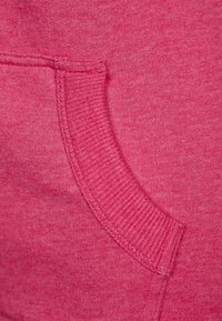 Primo piano del tessuto di una felpa rosa che presenta una tasca laterale con bordi a coste, una texture liscia e una distribuzione uniforme del colore.