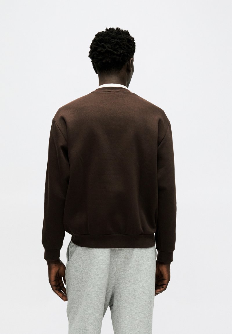 Persona con capelli corti e ricci vista di spalle, che indossa un maglione marrone scuro e pantaloni grigi chiari su uno sfondo chiaro e semplice.