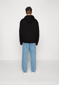 Felpa nera con cappuccio e maniche a costine, abbinata a jeans in denim baggy di un azzurro chiaro. L'outfit presenta un design minimalista e una texture morbida.