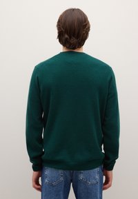 Dunkelgrüner Strickpullover mit langen Ärmeln und Rundhalsausschnitt, von hinten gezeigt, kombiniert mit hellblauen Jeans. Weiche Textur, schlichtes Design.