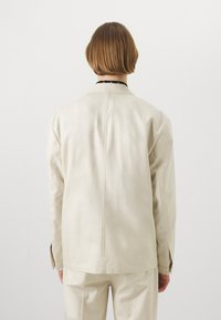 Beige läderjacka med en lös design, som har en söm bak, långa ärmar och krage. Enkel textur, minimalistisk stil.