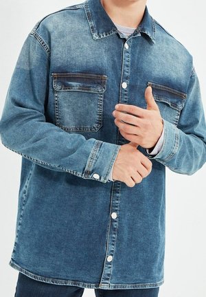 Mężczyzna poprawiający rękaw niebieskiej dżinsowej koszuli zapinanej na guziki z kieszeniami na piersi, stojący na białym tle.