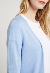 Helles blaues Cardigan aus weichem Strickmaterial, mit V-Ausschnitt und offener Vorderseite. Über einem weißen Oberteil getragen und zeigt eine entspannte Passform.