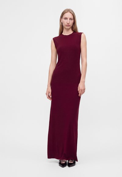Femme debout portant une robe bordeaux sans manches, longue jusqu'au sol, et des chaussures noires à bouts pointus sur un fond clair uni.