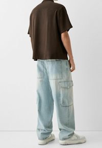 Osoba ubrana w luźne, jasnoniebieskie spodnie cargo z dużymi kieszeniami, białe trampki i koszulę z krótkim rękawem w ciemnym brązowym kolorze, stojąca na białym tle.