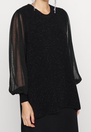 Haut sans manches noir scintillant à texture, superposé sur des manches longues transparentes et bouffantes, associé à des boucles d'oreilles pendantes en cristal.